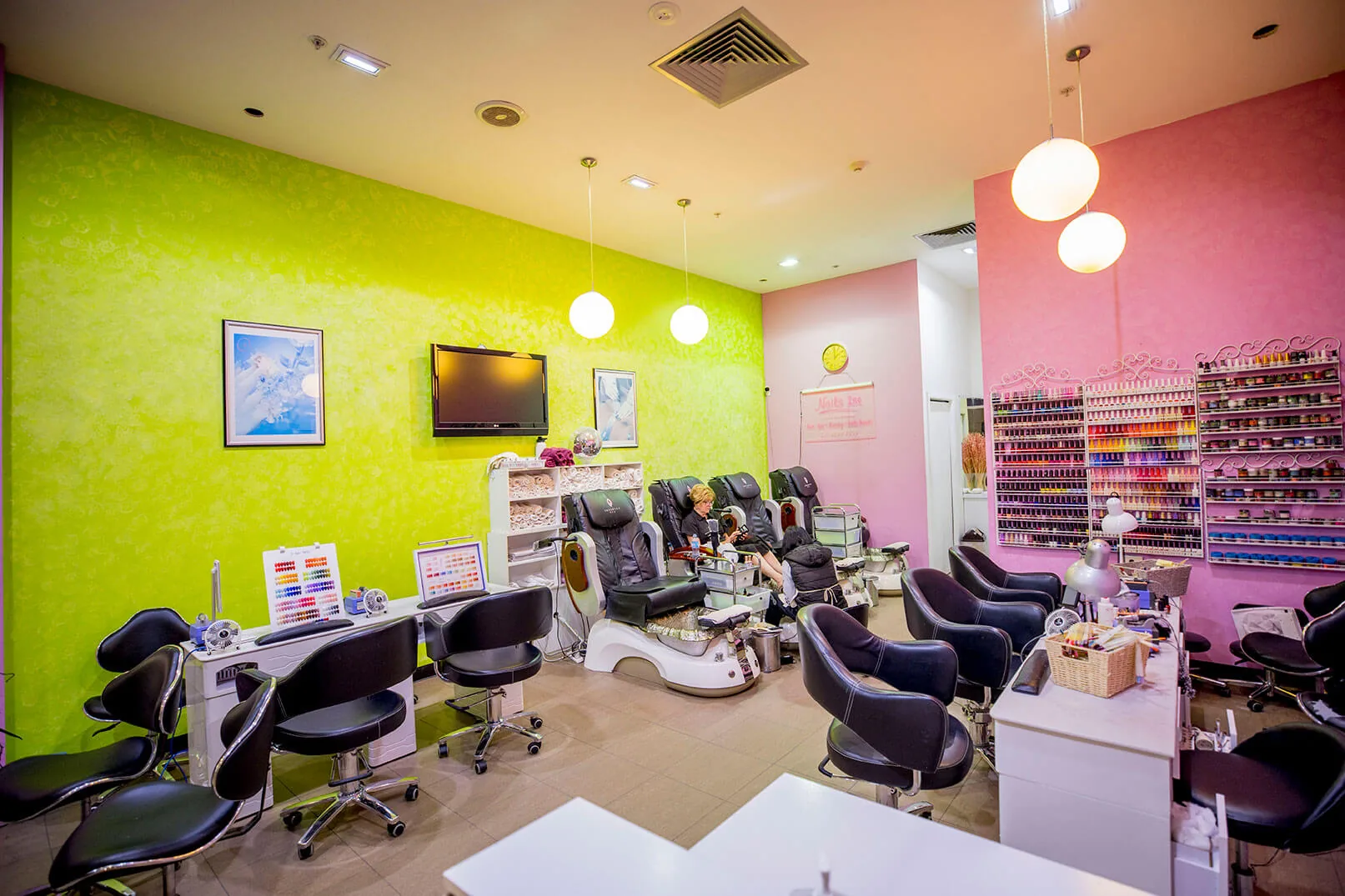 Wide view of the Nails 1st salon — lime-green feature wall, pedicure thrones, globe pendants and a technician at work