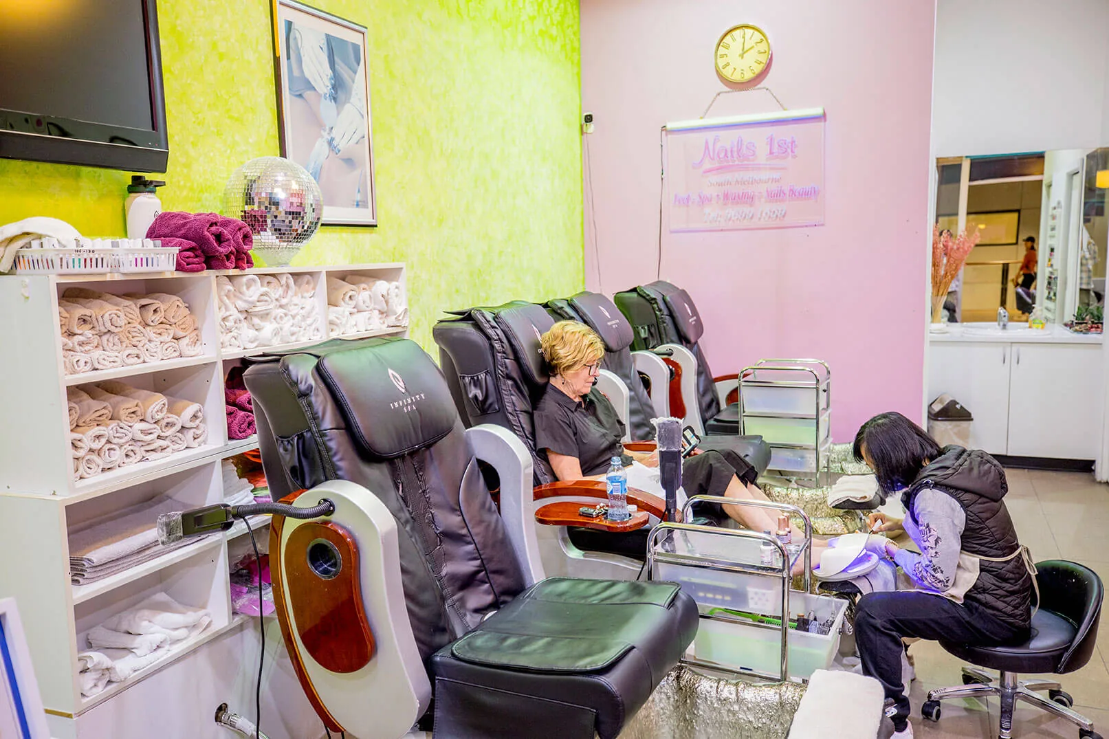 Row of pedicure thrones with a client and technician during treatment, Nails 1st wall banner visible