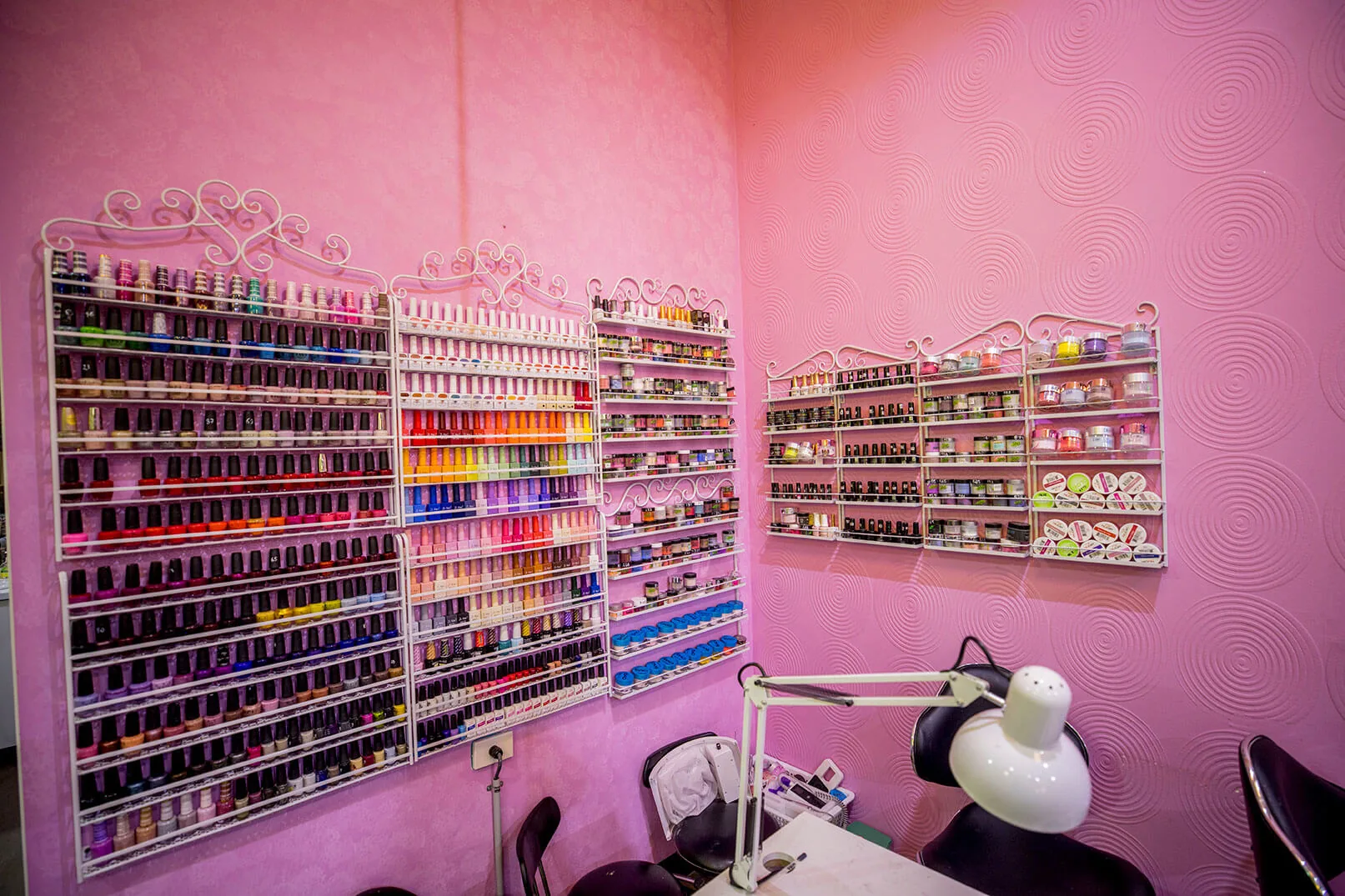 Manicure station framed by four wire racks of rainbow-sorted nail polish on pink walls